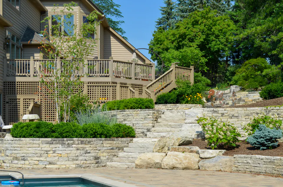 Poolside retaining wall below a sided house with deck stairs, paver deck, boxwood hedge, ornamental grasses, and planting.